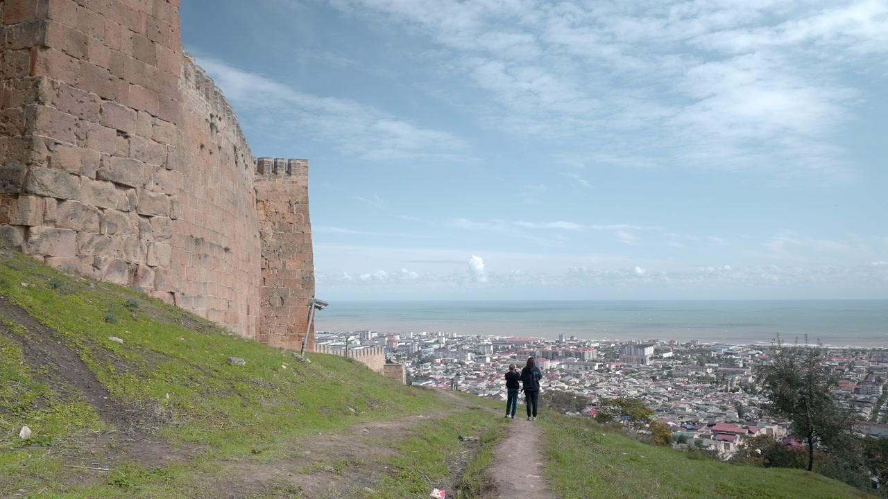 Two Figures Walking Fortress Path Above City, Stone Rampart On Left, Dirt Trail Leading Toward Sea Horizon. Creative