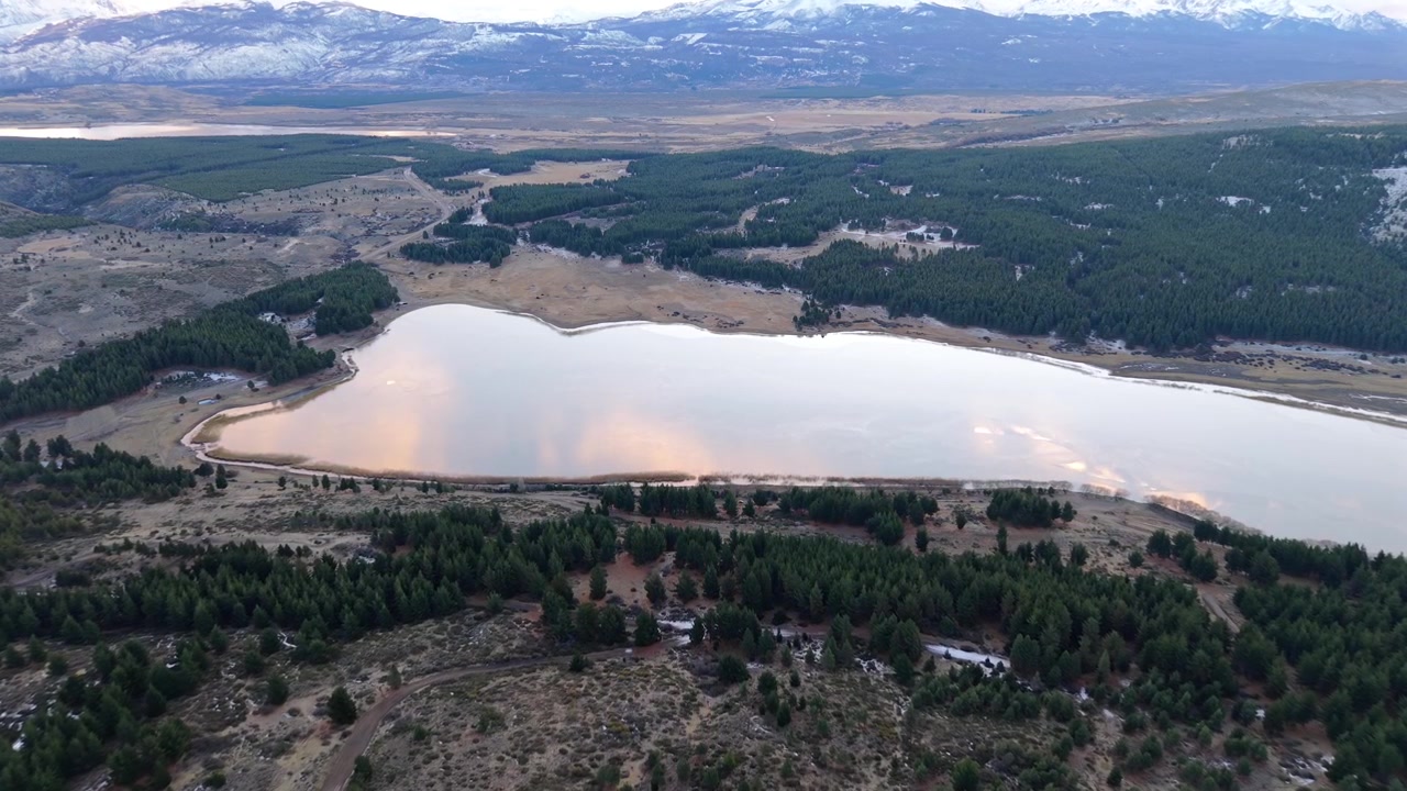 Un volo aereo si muove sopra foreste di pini verso una laguna calma che riflette il cielo al tramonto.