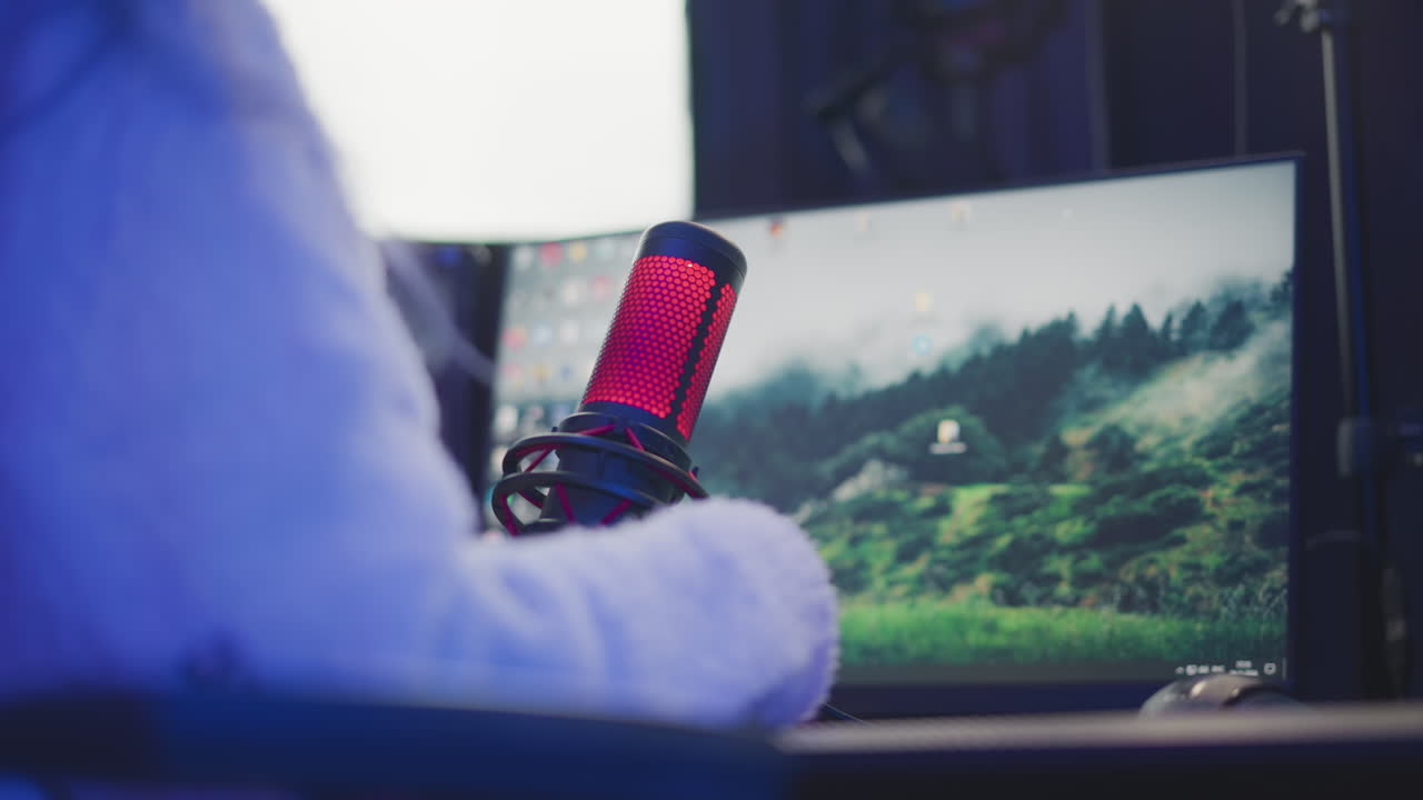 close up rear view of lady extending red condenser microphone toward herself while securing usb cable connection beside illuminated rgb light strip, speaker and keyboard on dark desk setup