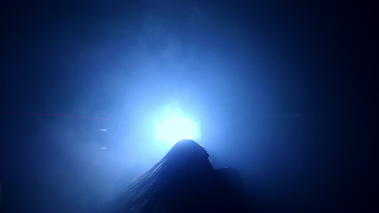 Long-haired female moving in front of the light. Silhouette of a dancer performing in the dark room with one lamp.