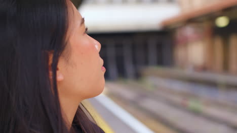 Close-Up-Of-Young-Woman-Waiting-On-Railway-Station-Platform-For-Train-Looking-At-Mobile-Phone