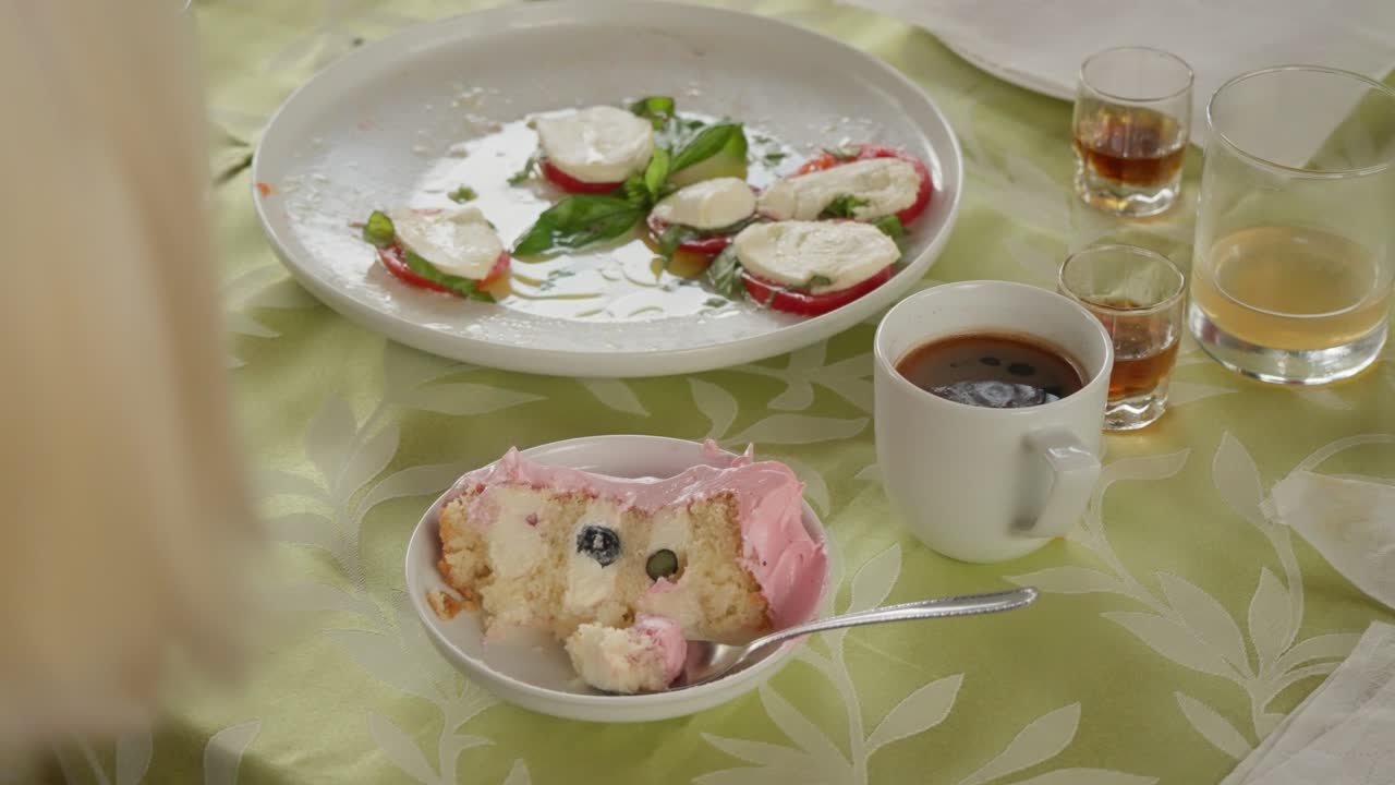 Primo piano di una tavola apparecchiata a festa con una fetta di torta alla crema con glassa rosa e mirtilli su un piatto, accanto a un'insalata caprese con pomodoro e mozzarella e una tazza di caffè nero a Vilnius.