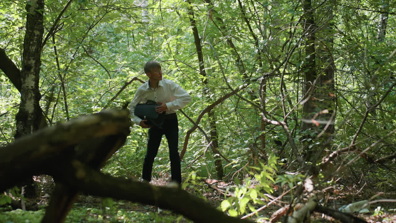 Scientific researcher in white shirt carrying backpack while bending carefully and walking through dense green forest under daylight, moving cautiously between trees and branches surrounded by lush