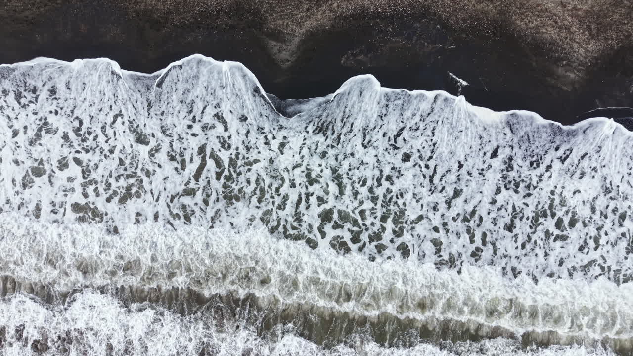 Onde si infrangono su una spiaggia di sabbia nera durante una giornata nuvolosa con acqua spumeggiante e una costa liscia