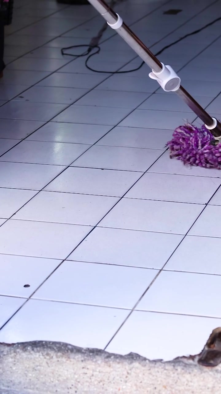 A person mops a tiled floor in a storefront, using a purple mop. Bright lighting highlights the clean environment