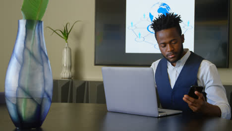 Front-view-of-young-black-businessman-talking-on-mobile-phone-while-working-on-laptop-in-office-4k