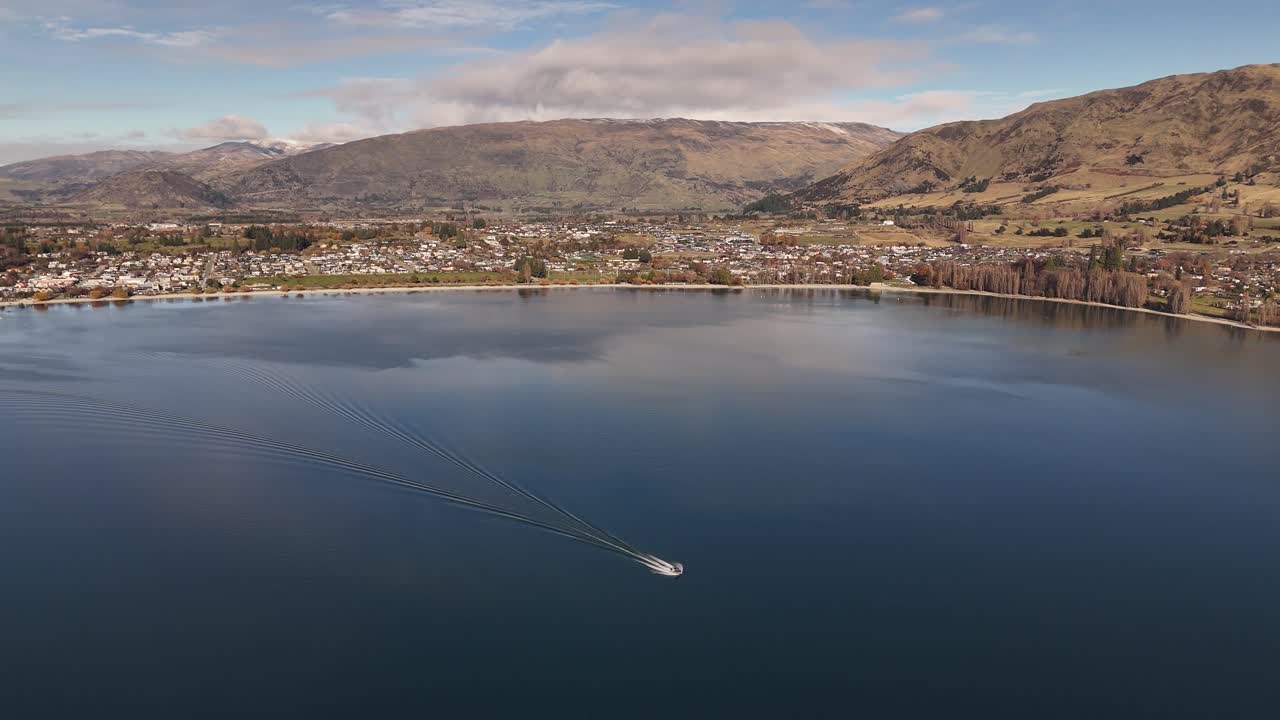 Aerial drone shot of motorboat cruising on calm Lake Wanaka with town and mountains in background at sunset, South Island, New Zealand
