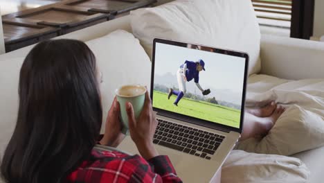 compuesto de una mujer sentada en casa sosteniendo un café viendo un juego de béisbol en una computadora portátil