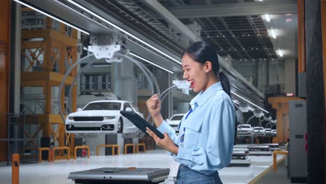 vista lateral de una trabajadora profesional asiática de pie con su tableta en una fábrica de automóviles ev, instalación industrial de alta tecnología, levanta el puño con una meta gritando después de comprobar en la tableta