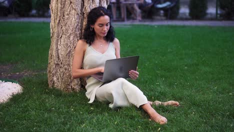 woman in dress sits below tree at the park on grass, holding laptop on her knees, typing, doing her work