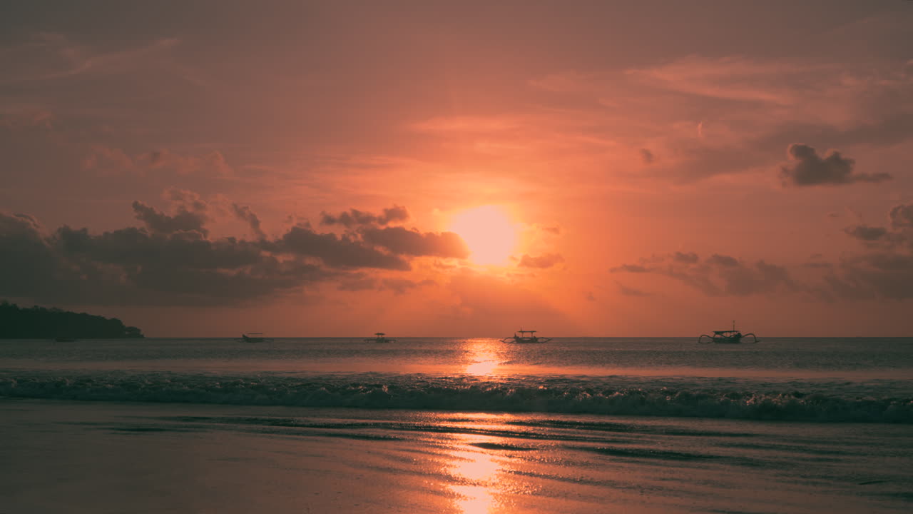 A breathtaking sunset paints the sky as boats gently float on the calm waters of Jimbaran Beach in Bali Indonesia as waves crash and roll on sandy shores