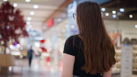 vista trasera de una mujer elegante con el cabello marrón largo caminando con confianza en un centro comercial, fondo borroso con tiendas iluminadas y compradores