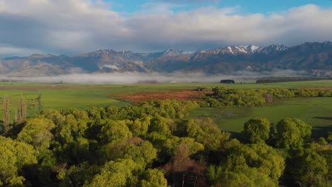 Slowly-flying-sideways-over-a-green-field-with-trees,-misty-mountain-range-in-the-background