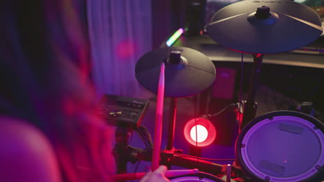 rear view of woman playing electronic drums under red and purple studio lights, striking cymbal and pads with drumsticks, drum module and cables visible, intimate indoor music practice atmosphere