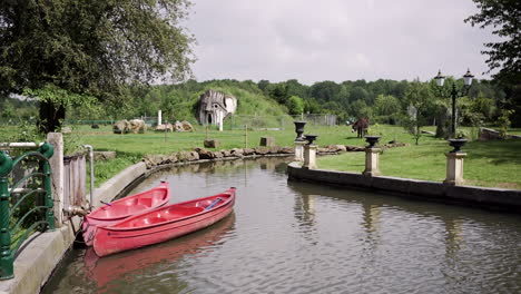 Peaceful-garden-scene-with-red-boats-docked-by-a-tranquil-lake,-surrounded-by-lush-greenery-and-grazing-animals-under-a-bright-sky