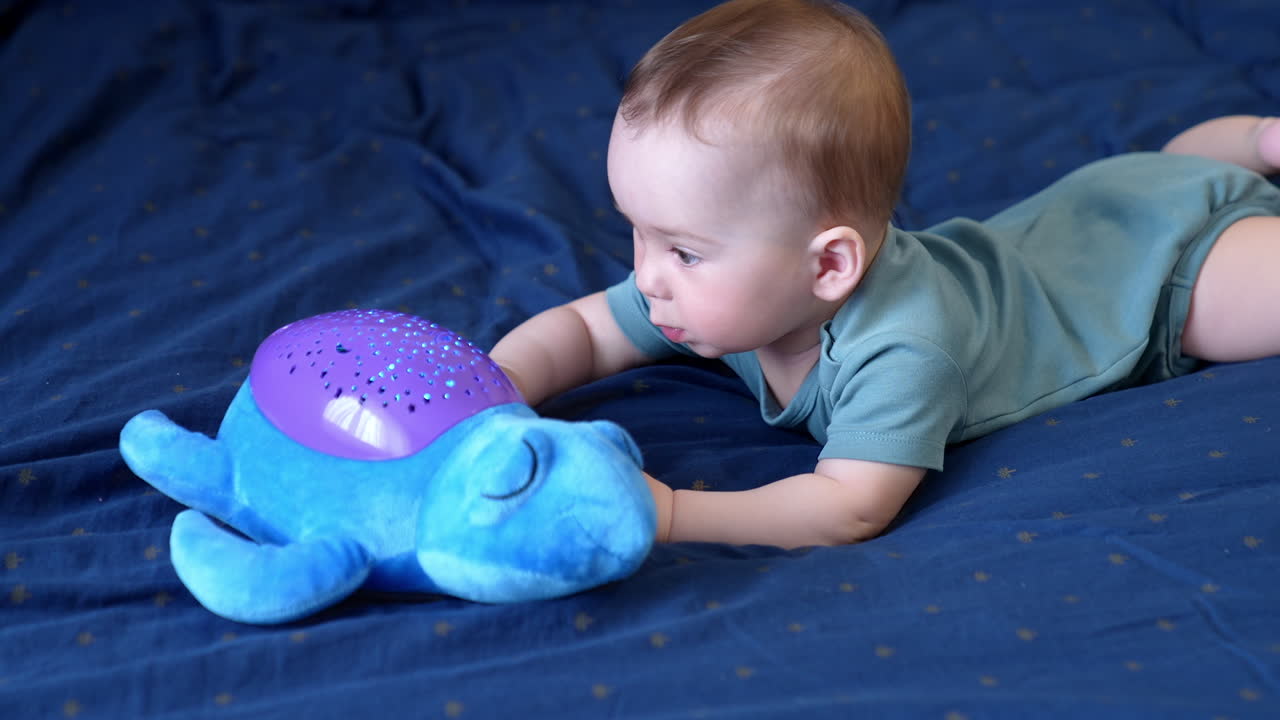 Cute lovely interested baby boy crawls to the toy lying on the bed in front of him. Caucasian baby playing with a soft toy on blue backdrop.