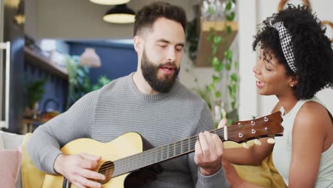 Video-of-happy-diverse-couple-relaxing-in-living-room,-man-playing-guitar-and-signing-to-woman