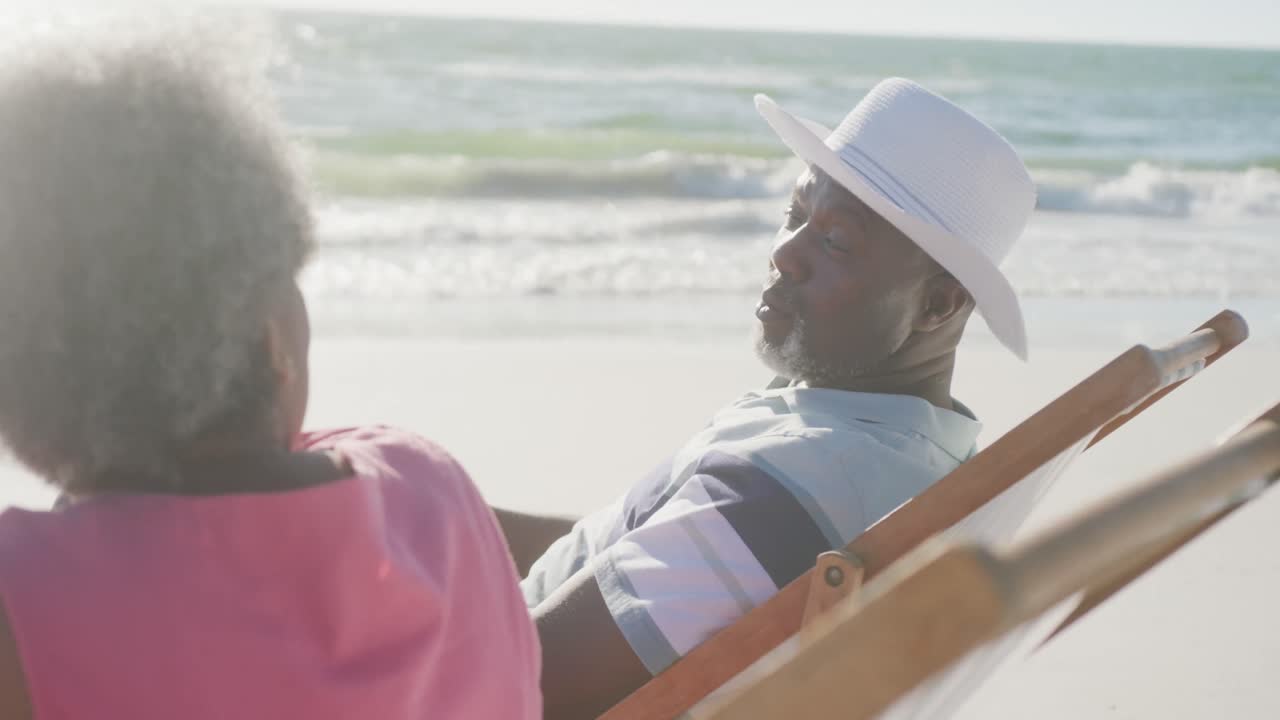 Happy senior african american couple sitting on deck chairs and talking at beach, in slow motion