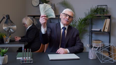 Smiling-senior-business-man-waving-with-stack-of-money-at-office-workplace-after-success-online-work