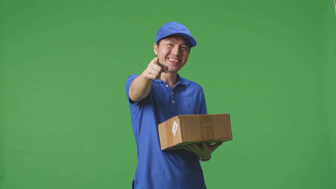 Corriere maschio asiatico in uniforme blu che sorride e fa un gesto di onestà mentre consegna un cartone su sfondo green screen in studio