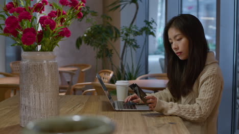 Young-pretty-Chinese-girl-works-on-computer-in-cafe,-checks-message-on-her-phone