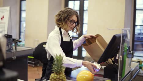 vendedora delgada y alegre con camisa blanca y delantal negro escaneando productos, frutas en el mostrador de caja en un supermercado brillante