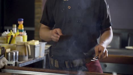 a chef prepares food on a hot grill with a spatula and tongs, creating smoke.