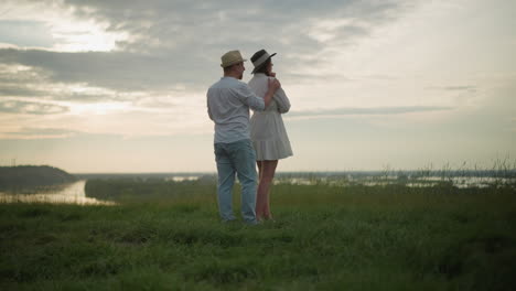un hombre con sombrero, camisa blanca y vaqueros camina hacia una mujer con un vestido blanco de pie en una colina cubierta de hierba al atardecer. la escena captura un momento sereno y contemplativo