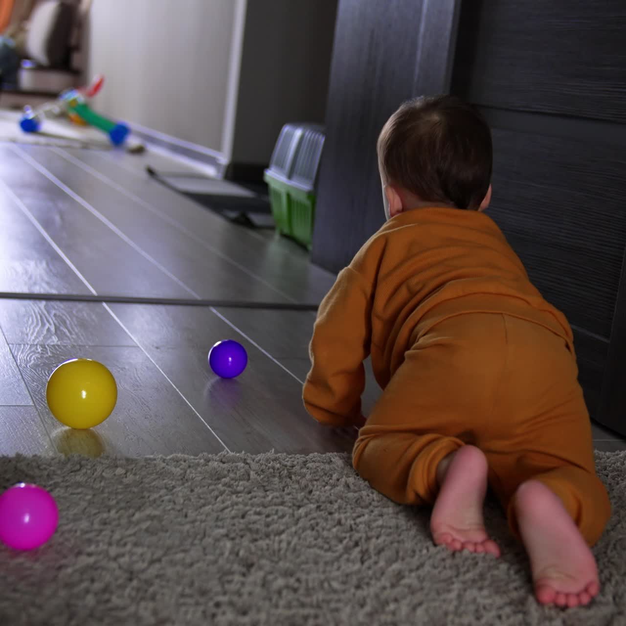 Little baby boy wearing orange sport suit crawls by the floor away from camera. Barefoot kid moves by the carpet towards the room door