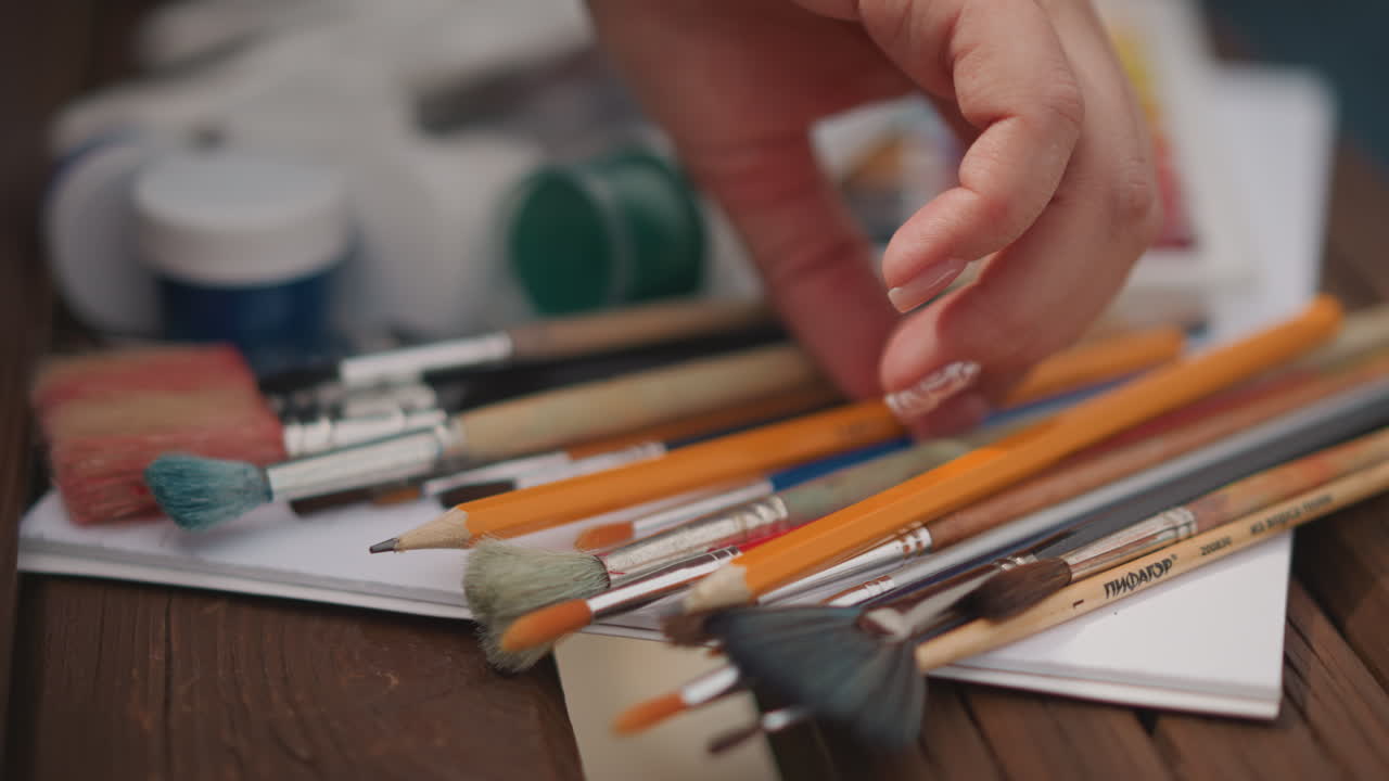 Closeup brushes hand selecting palette nearby. Soft studio light, jars paint and watercolor pans scattered on wooden table, gentle hand movement over bristles, careful selection of tools, tactile