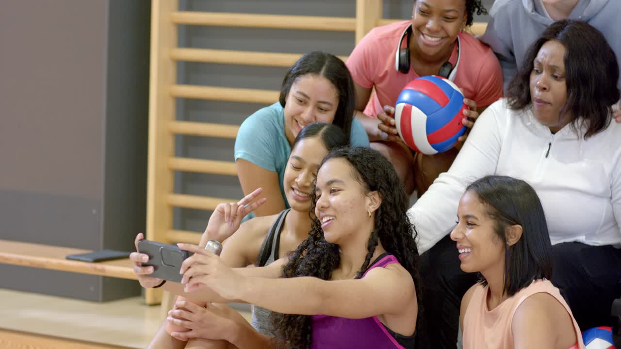 Weibliche Volleyball-Teamkolleginnen positionieren ihr Smartphone für ein Selfie, während sie auf der Tribüne einen Volleyball halten.