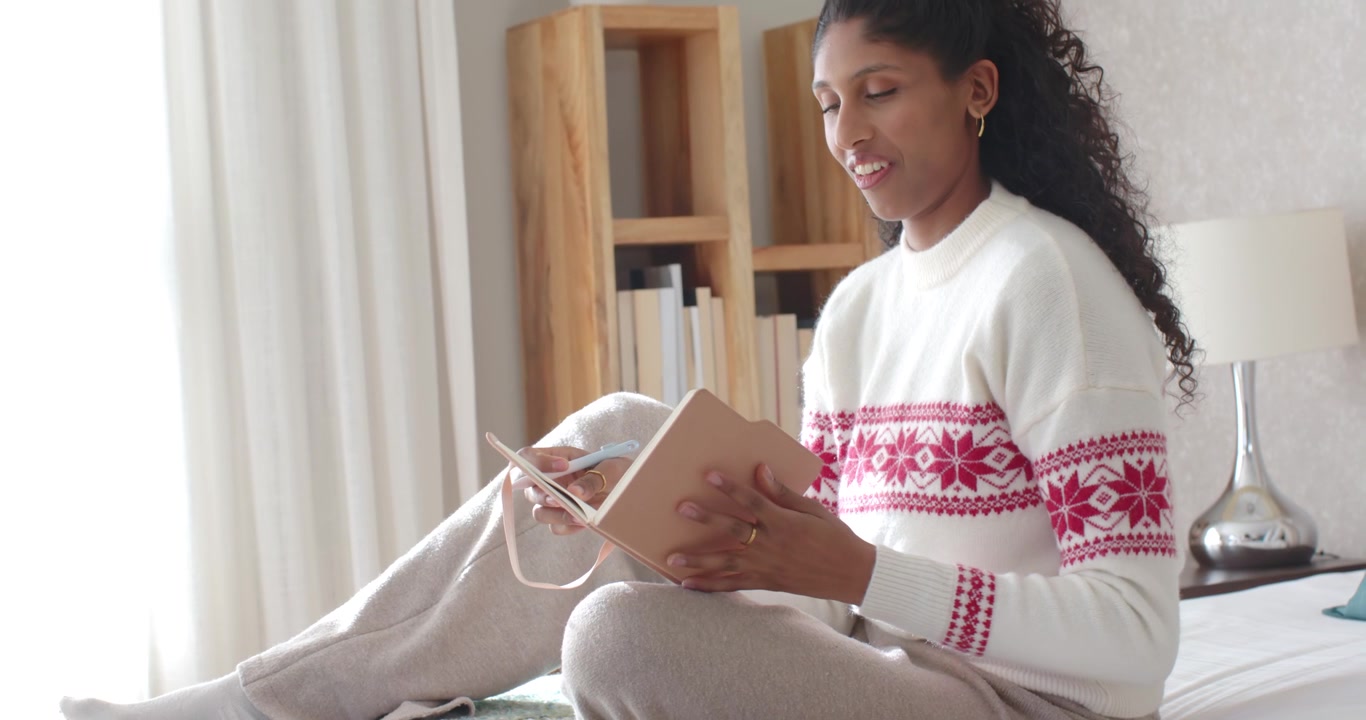 African American woman holding pink ribboned notebook flipping pages reading diary on bed by window