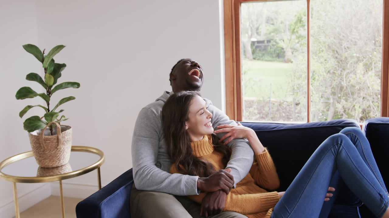 Happy diverse couple sitting on sofa laughing and embracing in home,copy space