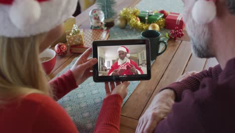 Pareja-Caucásica-Con-Gorros-De-Papá-Noel-Usando-Una-Tableta-Para-Una-Videollamada-Navideña-Con-Papá-Noel-En-La-Pantalla