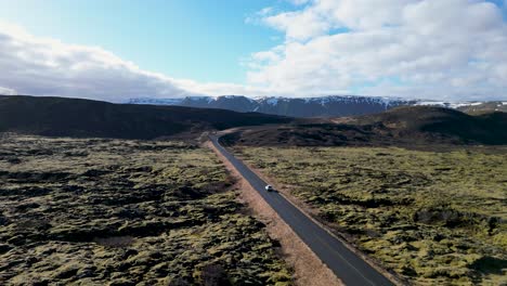 Imagínese-Volar-Sobre-El-Impresionante-Paisaje-De-Islandia,-Observando-Un-Automóvil-Que-Serpentea-Entre-Exuberante-Musgo-Y-Rocas-Negras,-Con-Montañas-Nevadas-En-La-Distancia-Bajo-Un-Cielo-Azul.