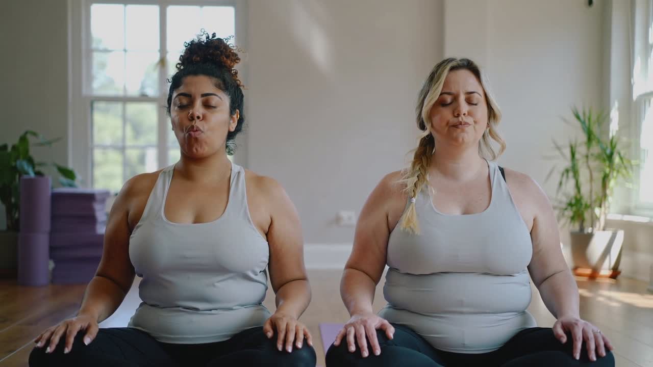 Su indicazione, due donne in canottiere grigie e leggings neri che praticano la respirazione ritmata su tappetini viola