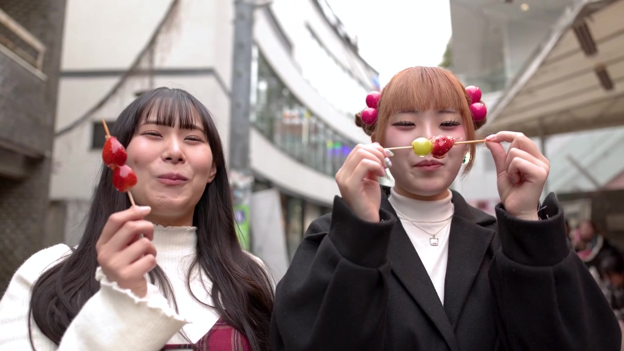 หญิงสาวสองคนกำลังกินผลไม้เสียบไม้เคลือบน้ำตาลบนถนนที่พลุกพล่านในโตเกียวช่วงกลางวัน