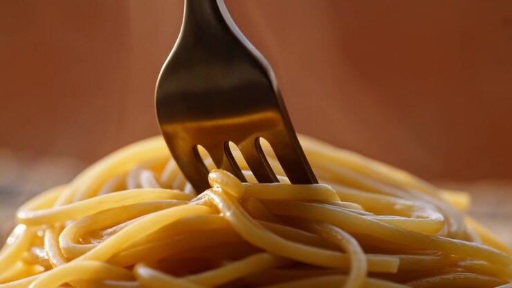 I rebbi scuri di una forchetta sollevano e raccolgono fili di spaghetti lucidi in una calda scena di cucina in primo piano, con un leggero movimento della pasta, sfocatura marrone tenue, riflessi dorati, texture ricca e un'appetitosa atmosfera rustica.