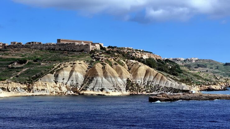 Suggestiva vista estiva di Fort Chambray a Gozo, Malta, che si affaccia su ripide scogliere di argilla. La scena, caratterizzata da toni terrosi e un mare blu intenso, è stata ripresa da una prospettiva dal basso di una barca sull'acqua durante una giornata di sole.