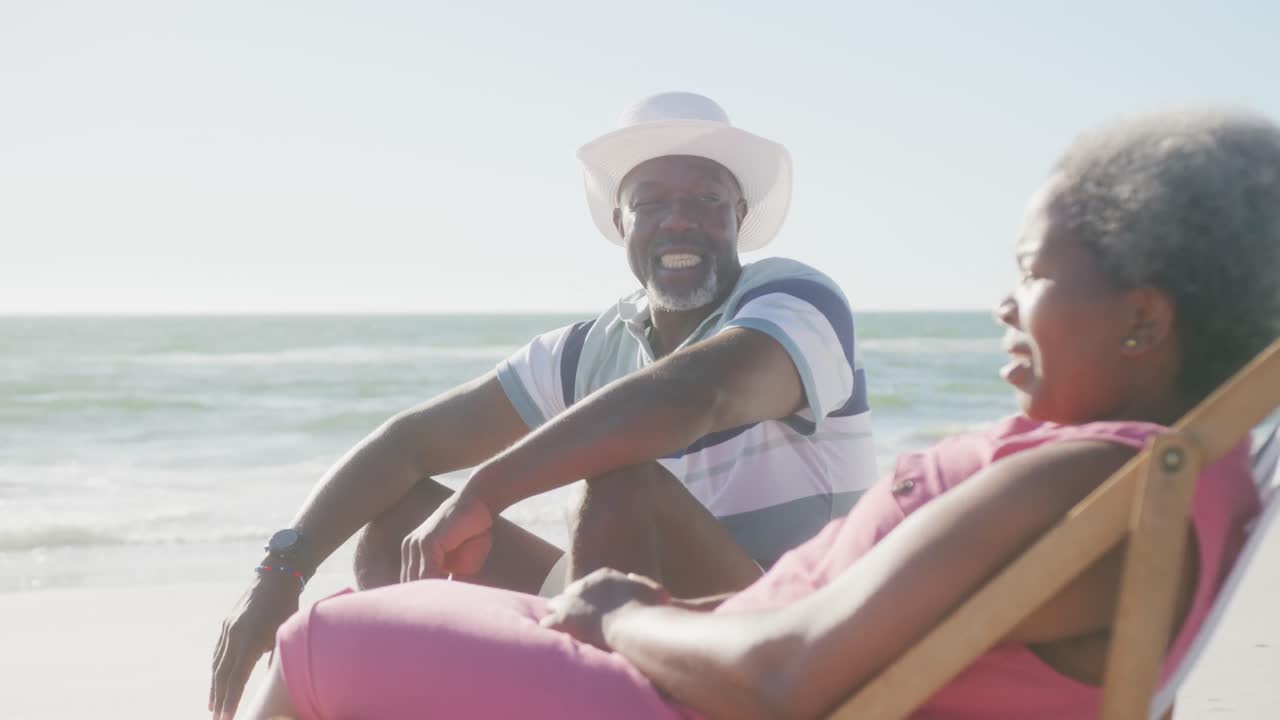 Happy senior african american couple sitting on deck chairs and talking at beach, in slow motion