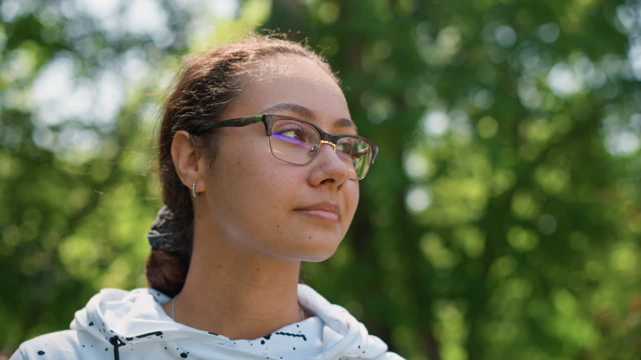 Mujer birracial ajustándose la sudadera con capucha en un parque con gafas y smartwatch, la luz del sol filtrándose entre los árboles, expresión tranquila y contemplativa, preparándose para un paseo al aire libre o ejercicio suave, bokeh suave y frondoso