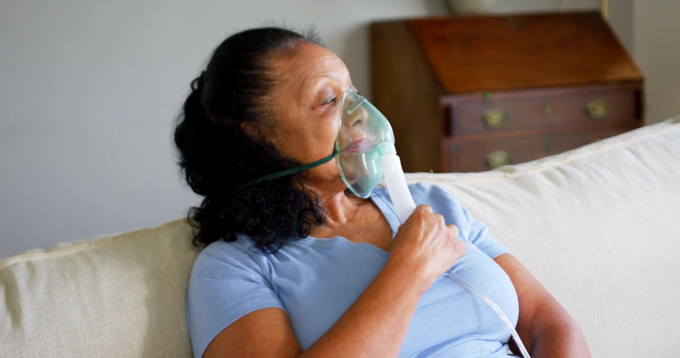 Mulher afro-americana idosa usando nebulizador no sofá em casa, segurando máscara e tubos para terapia.