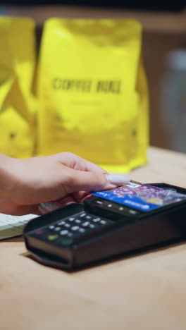 primer plano de una persona con un traje vintage sosteniendo una tarjeta sobre un lector de tarjetas para hacer el pago, con la muñeca adornada con pulseras y un reloj, bolsas de café amarillas y papeles de menú son visibles en la mesa