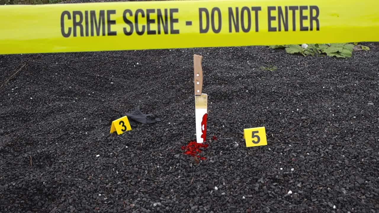 Police yellow colored line or crime scene tape in front of yellow crime markers that are placed besides murder evidence like bloody kitchen knife and a nylon black glove on gravel ground outdoors