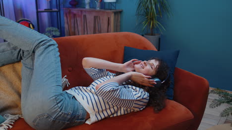 Happy young woman in wireless headphones relaxing sitting on sofa at home listening favorite music