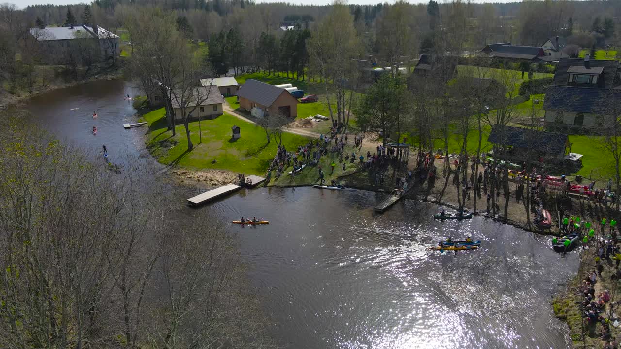 High static aerial view of paddle boats reach the shoreline for a break. Kayaks landing and exiting on shore. People gathered on the riverbank to support teams at Vohandu Marathon, rowing competition