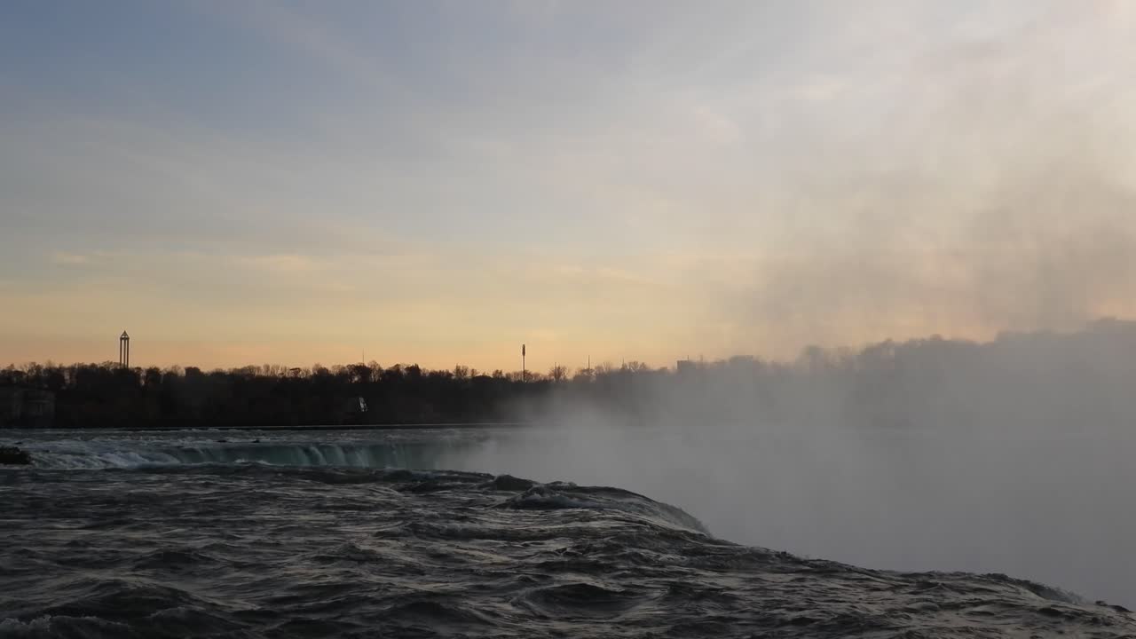 Aerial drone footage captures sunrise over Niagara Falls Canada as mist gently rises into golden sky with calm flowing water reflecting soft light near edge of powerful natural wonder