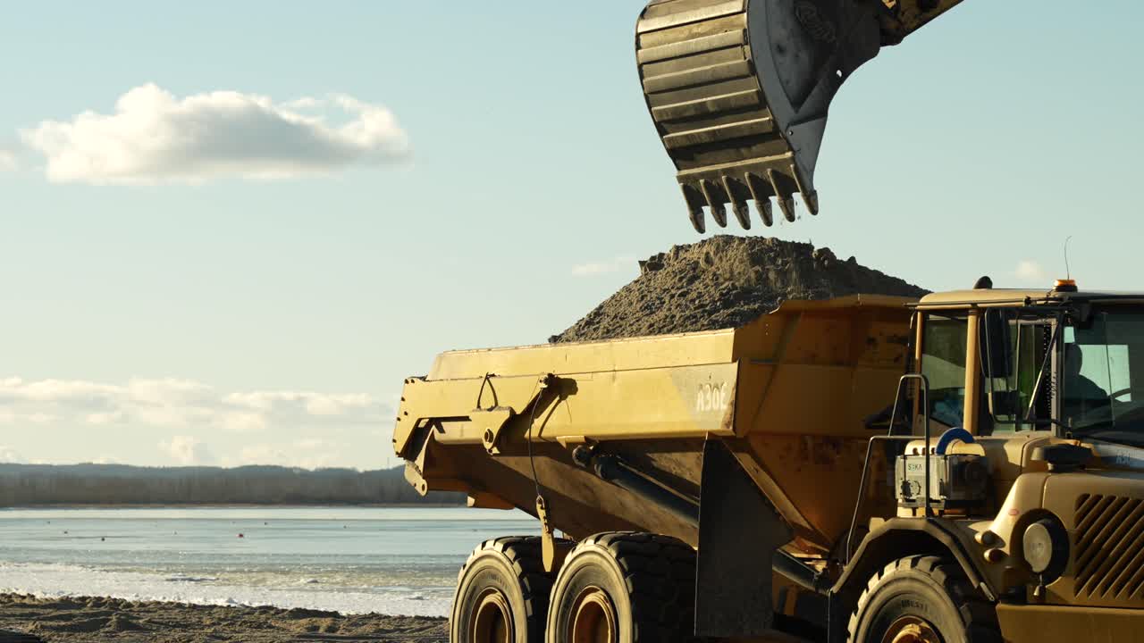 Excavator loading industrial dump truck at beach building site, slow motion