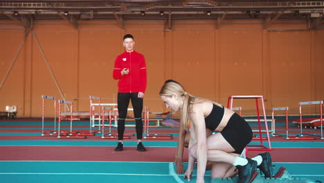 dos atletas multiétnicas tomando posición en la línea de salida y luego corriendo en una pista cubierta mientras un entrenador masculino mide el tiempo con un cronómetro