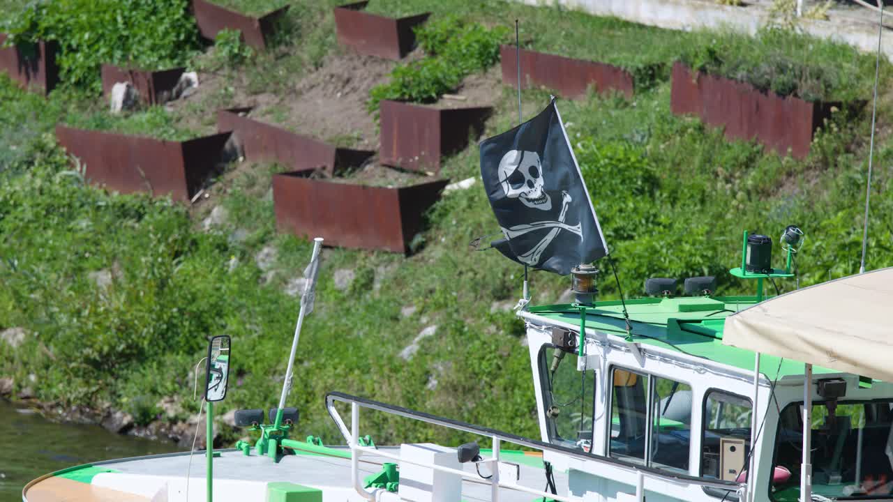 Pirate flag waves atop green-roofed boat docked riverside, bright daylight, Prague, steady camera